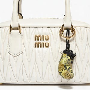 May include: Cream-coloured quilted leather handbag with gold-tone hardware and a top handle. The bag features the brand's logo in gold lettering. A small, decorative charm with a snakeskin pattern hangs from the handle.