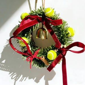 以下が含まれることがあります： 緑色のベースに、黄色のテニスボール、赤いテニスラケット、金のベルをあしらった装飾的なリース。 リースは深紅のリボンで飾られており、お祝いの装飾に適しています。
