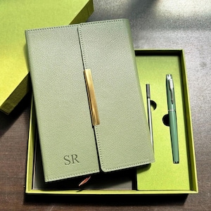 Peut inclure: Un journal en cuir vert sauge avec un fermoir en métal doré, présenté dans un coffret cadeau assorti. Le journal est estampillé des initiales "SR". Un stylo et une recharge assortis sont inclus dans la boîte.