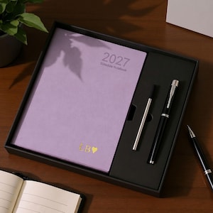 May include: A lavender 2027 schedule notebook with gold embossed initials LB and a heart icon, presented in a black gift box with a silver pen and a black fountain pen. An additional black fountain pen rests outside the box on the surface.