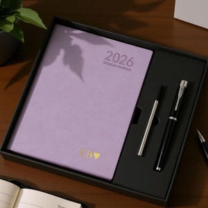 May include: A lavender 2026 schedule notebook with gold lettering and a black pen set in a black box. The notebook has the initials "LB" and a heart symbol. The pen set includes a black fountain pen and a silver pen.