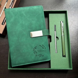 Könnte beinhalten: Ein grünes Notizbuch- und Stift-Set in einer Geschenkbox. Das Notizbuch hat einen silbernen Verschluss und den Namen "Maisie" mit einem Lorbeerkranz-Design. Der Stift und eine Mine befinden sich in einer passenden grünen Box.