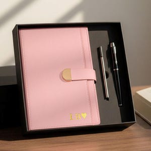 May include: A pink leather journal with a gold clasp and the initials "LB" with a heart symbol, presented in a black gift box. Includes a silver pen and a black pen. The journal is approximately 20 cm tall.