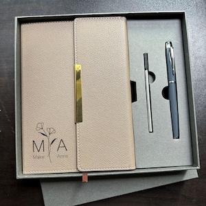 Peut inclure: Un ensemble de journal et de stylo en cuir beige dans une boîte grise. Le journal a un fermoir doré et le texte "MYA Make Anne" avec un motif floral. L'ensemble comprend une recharge de stylo argentée et un stylo gris.