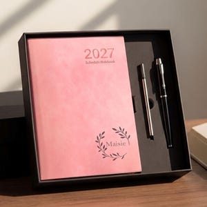 May include: A pink 2027 schedule notebook with a leaf wreath design and the name Maisie printed on the cover, presented in a black gift box alongside a black ballpoint pen and a thin silver metal pen refill in a custom-fitted black insert.