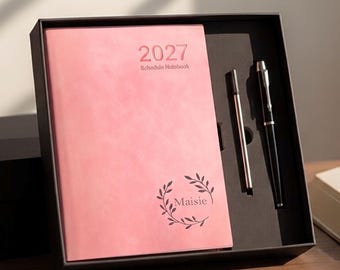 May include: A pink 2027 schedule notebook with a leaf wreath design and the name Maisie printed on the cover, presented in a black gift box alongside a black ballpoint pen and a thin silver metal pen refill in a custom-fitted black insert.
