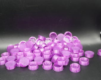 May include: A pile of small, round, purple plastic objects. Each object has a flat, circular top and a cylindrical base. The objects are scattered on a dark surface, suggesting a craft or DIY project.