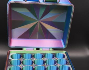 Könnte beinhalten: Eine offene, schillernde Aufbewahrungsbox mit einem geometrischen Design auf der Innenseite des Deckels und einem Raster aus kreisförmigen Fächern im Boden. Die Box ist hauptsächlich grün und blau mit Hauch von Lila und Rosa.