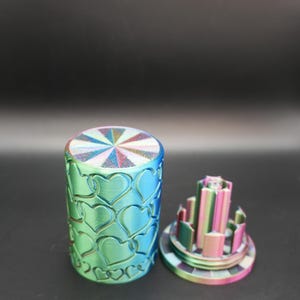 May include: A cylindrical container with a lid, both featuring a heart pattern. The container is a gradient of blue and green, with a rainbow-colored top. The lid has a multi-colored, architectural design.