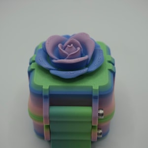 May include: A square, multi-colored 3D printed box with a gradient finish of green, blue, and pink. The lid features a prominent, sculpted rose in shades of purple and blue. The box includes a hinged side mechanism secured with two silver metal screws.