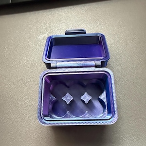 May include: A rectangular, 3D-printed storage box with a hinged lid, featuring a vibrant iridescent purple and blue color gradient. The interior base contains a molded insert with six circular indentations and two central diamond-shaped posts.