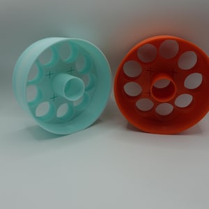 May include: Two 3D-printed objects. One is a light blue cylinder with evenly spaced holes and a central opening. The other is an orange cylinder with similar features. Both have cross-shaped markings. The objects are likely molds or tools.