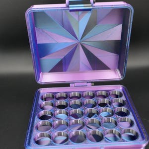 May include: Open, purple, geometric-patterned box with a hinged lid. The interior features a grid of circular compartments. The lid's interior has a starburst design in shades of purple, blue, and black. The box is on a black surface.