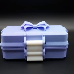 May include: A light purple box with a bow on top. The box has a white clasp and is rectangular. The bow is the same color as the box. The box is on a black surface.