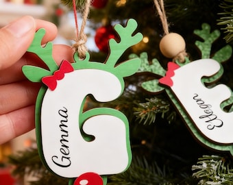 May include: A wooden Christmas ornament shaped like the letter G, painted white with green reindeer antlers, a red bow, and a red nose. The name Gemma is printed in black script on the letter. A second ornament shaped like the letter E is visible.