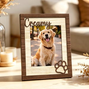 May include: A rectangular wooden picture frame with a dark brown border and a light wood-grain inner frame. The frame features the word Creamy in dark script at the top and a dark brown paw print cutout at the bottom right corner.