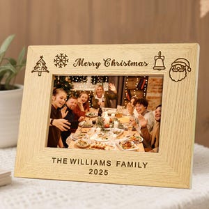 Puede incluir: Un marco de fotos de madera clara con las palabras "Merry Christmas" y "THE WILLIAMS FAMILY 2025" grabadas. El marco contiene una foto familiar. También están grabados iconos de árbol de Navidad, copo de nieve, campana y Papá Noel.