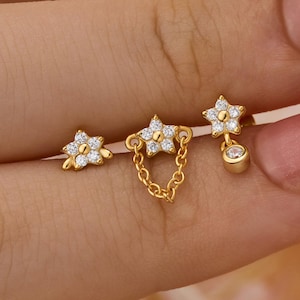 May include: A set of three gold earrings. One is a small flower stud, another has two flower studs connected by a chain, and the third has a flower stud with a dangling charm. All earrings have small, clear stones.