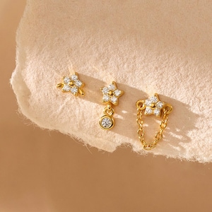May include: Three gold earrings with floral and star designs. One earring features a small flower with a cluster of clear stones. Another has a dangling star with a single clear stone. The third earring has a star connected to a chain.