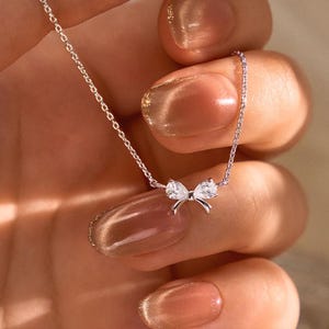 May include: A silver necklace with a bow pendant, set with two clear gemstones. The necklace is held in a hand, highlighting the fine chain and the bow's detailed design. The jewellery is elegant and suitable for various occasions, with a subtle sparkle.