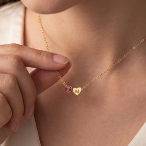 May include: A delicate gold necklace featuring a small heart pendant with the letter "M" engraved on it, and a round pink gemstone. The necklace is worn around the neck.
