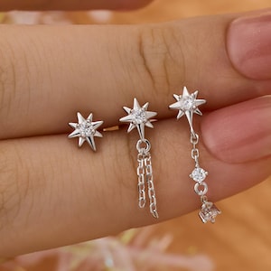 May include: Three silver star-shaped earrings with clear gemstones. One is a stud, another has a chain, and the third has a dangling design. The earrings are displayed against a warm, peachy background.