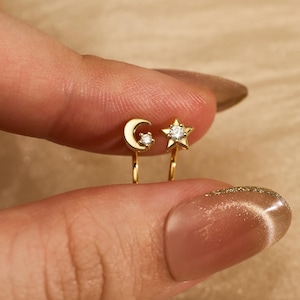May include: A pair of gold-coloured clip-on earrings, one shaped like a crescent moon and the other a five-pointed star. Both earrings feature a small, clear gemstone. The earrings are held between fingers against a soft, neutral background.