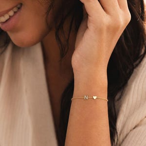 Könnte beinhalten: Ein goldfarbenes Armband mit der Buchstaben-Initialie "N" und einem Herzanhänger. Das filigrane Armband befindet sich am Handgelenk einer Person. Der Text "Dear Letter" ist unten im Bild zu sehen.