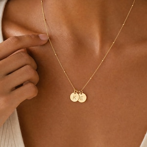 May include: A gold-toned necklace featuring a delicate chain with small evenly spaced beads and two circular pendants. Each pendant is engraved with a single uppercase letter, specifically K and E, hanging from the centre of the chain.