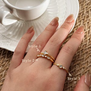 May include: Three gold rings are shown on a hand. Style 1 has a marquise-cut gemstone, Style 2 is a double band, and Style 3 has a small round stone. The rings are on a hand with long nails, near a white teacup and plate. The text "Dear Letter" is visible.