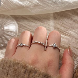 May include: Three silver rings are shown on fingers. One ring has a central round stone with smaller stones, another has a row of small stones, and the third has a marquise-shaped stone. The rings are on a hand with a neutral-toned sweater. The rings are delicate.