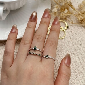 May include: Three silver rings with green gemstones and small white stones are displayed on a hand. The rings feature various designs, including an oval green stone and a band with multiple small green stones. The hand has long, polished nails.