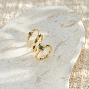 May include: Three gold rings featuring green gemstones and small white stones. The rings are presented on a white, textured surface. One ring has a twisted band, the others have smooth bands.