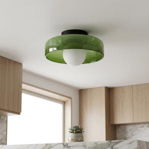 May include: A contemporary ceiling light, featuring a green, bowl-shaped shade and a white globe bulb. The light fitting has a black base and is set against a white ceiling. Wooden cabinets and a window are visible in the background.
