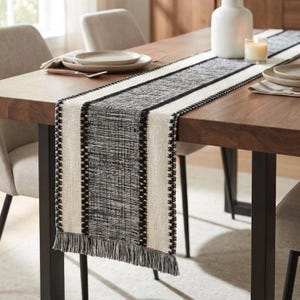 Peut inclure: Chemin de table tissé avec un motif texturé noir et blanc. Le centre est gris, bordé de rayures crème et d'une bordure noire. Des franges ornent les extrémités. Le chemin de table est présenté sur une table en bois.
