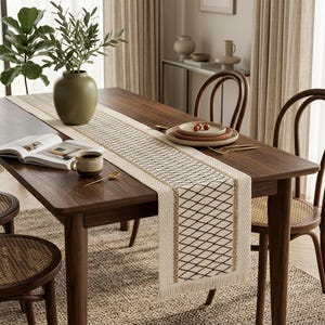 Peut inclure: Une table à manger en bois avec un chemin de table tissé à motif de losanges géométriques. La table est dressée avec des assiettes, une tasse à café et un vase avec de la verdure. Les chaises ont des assises tissées.