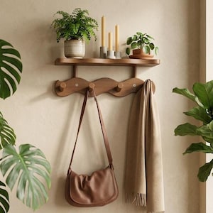 Puede incluir: Una estantería de pared de madera con un perchero ondulado debajo. La estantería contiene plantas en macetas y velas. Un bolso de hombro de cuero marrón cuelga del perchero, junto con una bufanda beige. La pared es de color neutro.