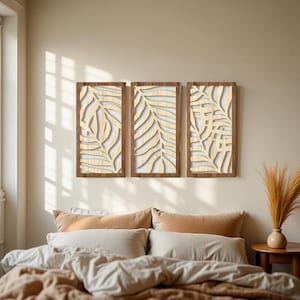 Peut inclure: Trois œuvres d'art murales rectangulaires en bois avec des motifs de feuilles. Chaque pièce a un cadre marron foncé et un motif de feuilles découpées de couleur claire. L'art est monté sur un mur de couleur neutre au-dessus d'un lit avec une literie de couleur neutre.
