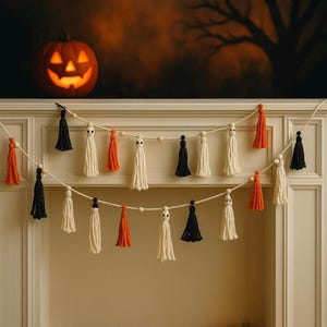 Peut inclure: Guirlande de pompons d'Halloween avec des pompons noirs, orange et crème, enfilés avec des perles en bois. Une citrouille sculptée avec un visage lumineux est en arrière-plan, créant une ambiance automnale.