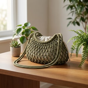 Peut inclure: Un sac à bandoulière en macramé vert avec un intérieur blanc à cordon de serrage est posé sur une table en bois. Le sac a un motif en filet tissé et de longues anses en corde. Des plantes en pot sont en arrière-plan.