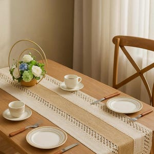Peut inclure: Une table en bois dressée pour un repas, avec un chemin de table en dentelle et toile de jute crème. La table est ornée d'assiettes, de tasses et de couverts blancs. Un arrangement floral doré ajoute une touche décorative.