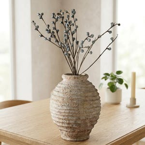 Peut inclure: Un vase en céramique beige texturé et arrondi avec des rainures horizontales, contenant plusieurs branches fines brun foncé ornées de petits bourgeons métalliques argentés en forme de cloche s'étendant vers le haut.