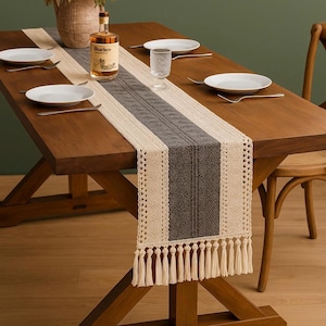 Peut inclure: Une table à manger en bois avec un chemin de table décoratif, des assiettes blanches et des couverts. Le chemin de table est beige, gris et crème avec des franges. Une bouteille de liqueur et un verre sont également sur la table.