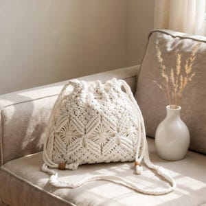 Może przedstawiać: Kremowa torba ze sznurkiem z makramy z geometrycznym wzorem. Torba ma długi, pleciony pasek i drewniane koraliki. Leży na beżowej sofie obok białego wazonu z suszonymi kwiatami.