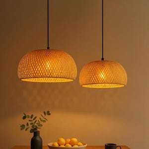 May include: Two woven pendant lights with dome-shaped shades hang from black cords. The shades are a warm, golden colour, and the woven pattern allows light to filter through. The lights are suspended above a table with a vase, bowl of fruit, and a mug.