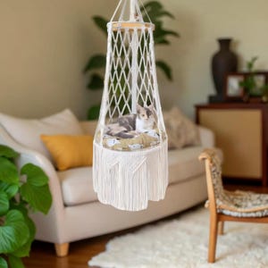 May include: A white macrame cat bed with a round, patterned cushion. The cat bed is suspended from the ceiling and has a fringed base. A grey and white cat is resting inside the bed.
