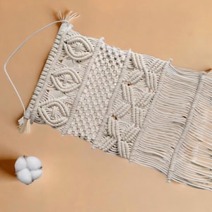 May include: Cream-colored macrame wall hanging with intricate woven patterns. The design features geometric shapes, knots, and fringe. A cotton boll is placed near the wall hanging. The background is a solid, light tan color.