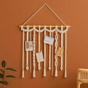 Puede incluir: Un expositor de fotos de macramé color crema con una clavija de madera y un triángulo colgante. La pantalla presenta cordones trenzados y rectos con borlas y cuentas de madera. Varias fotos pequeñas están sujetas a los cordones.