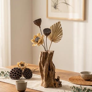 Può includere: Un vaso rustico in legno contiene baccelli di loto secchi, una foglia a forma di ventaglio e un delicato fiore giallo. Il vaso è posto su un tavolo di legno con altri elementi decorativi, creando un'estetica naturale e terrosa. Il vaso è alto circa 25 cm.