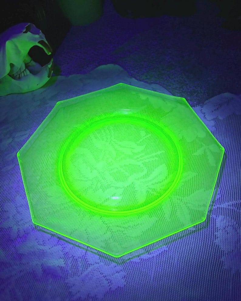 May include: A glowing green octagonal plate with a floral pattern. The plate is made of glass and has a raised rim. The plate is on a white and blue surface. A skull is in the background.
